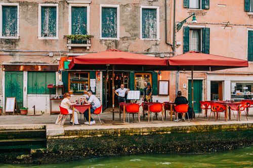 Free Charming outdoor café by a Venetian canal, featuring vibrant red chairs and patrons enjoying a relaxed day. Stock Photo