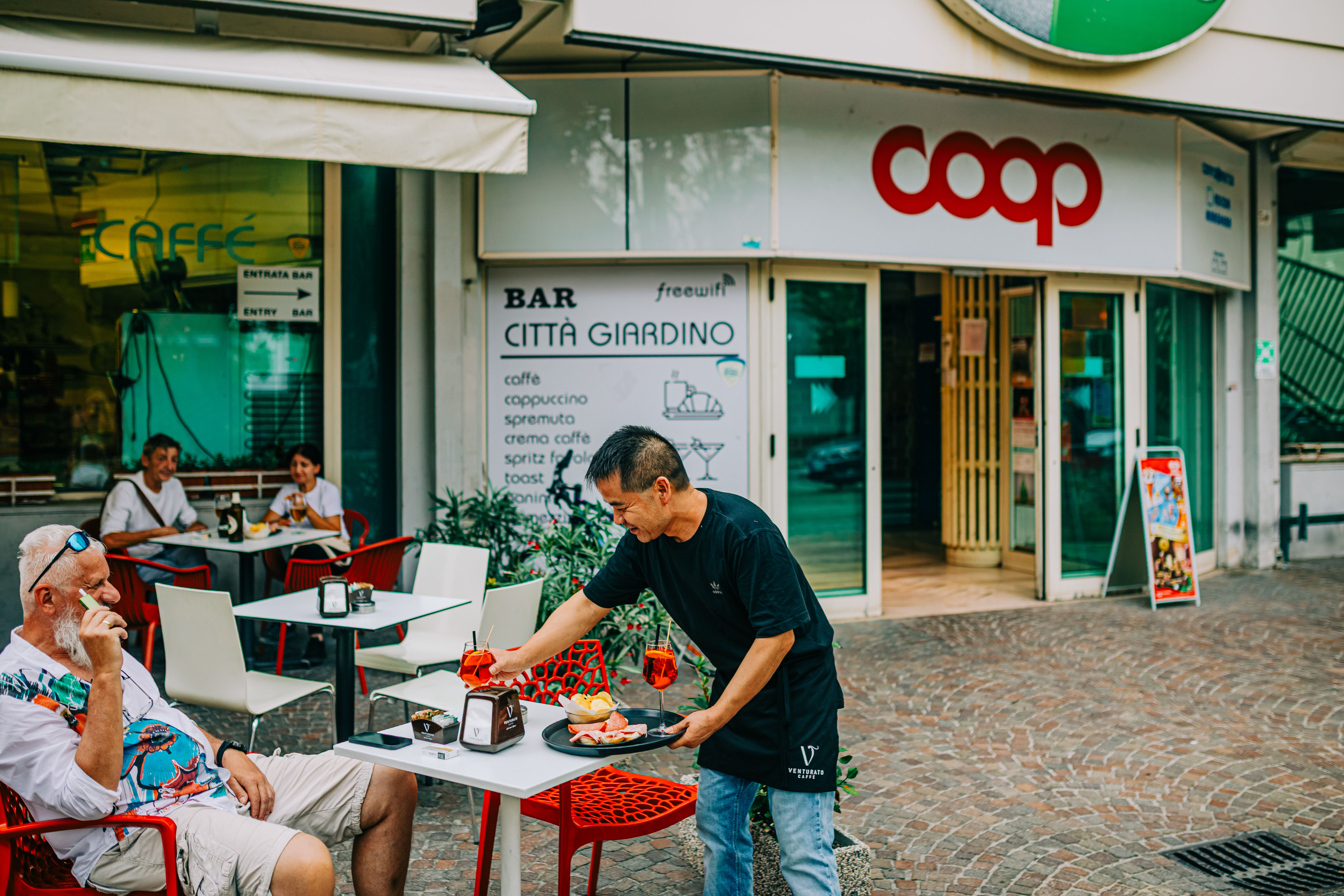 A bustling outdoor cafe with a waiter serving drinks and patrons enjoying snacks.