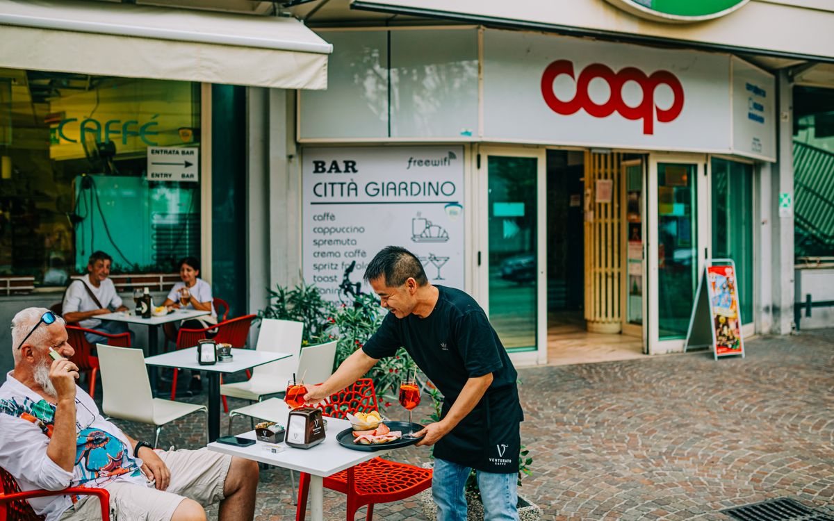 A bustling outdoor cafe with a waiter serving drinks and patrons enjoying snacks.