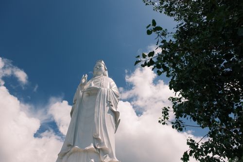 Free A serene white statue stands tall under a clear blue sky with surrounding trees. Stock Photo