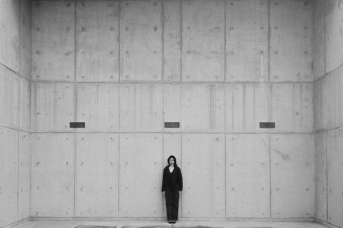 Free A woman stands alone in a stark, minimalist concrete room, embodying solitude and modern design. Stock Photo