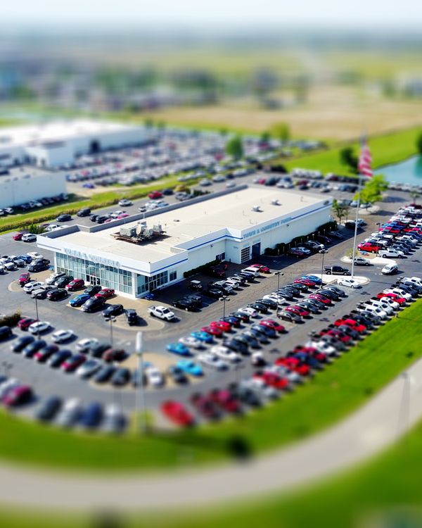 Aerial high-angle view of a bustling car dealership surrounded by parked cars in a green landscape.