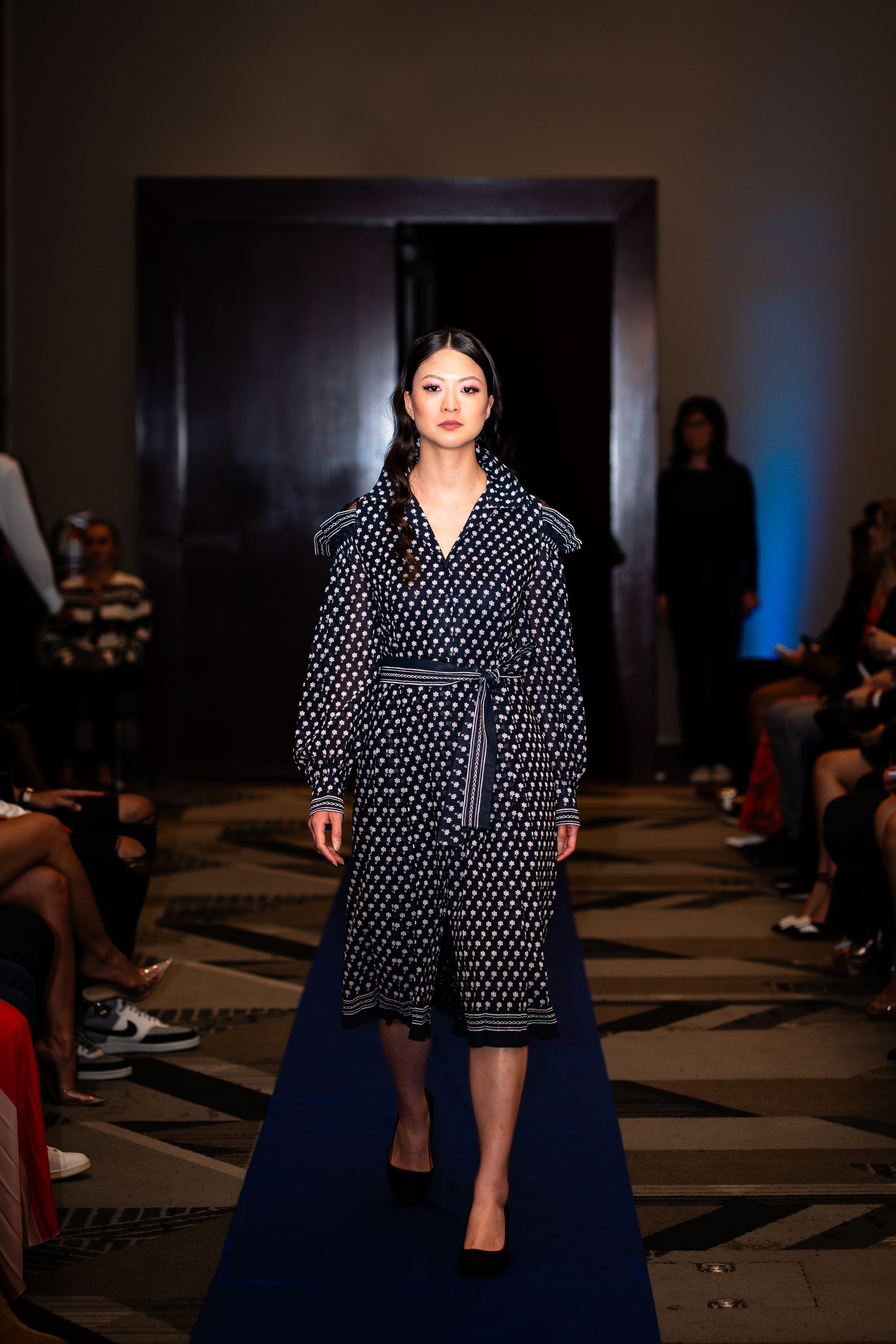 Modelo com um elegante vestido de bolinhas desfila na passarela durante evento de moda em local fechado.