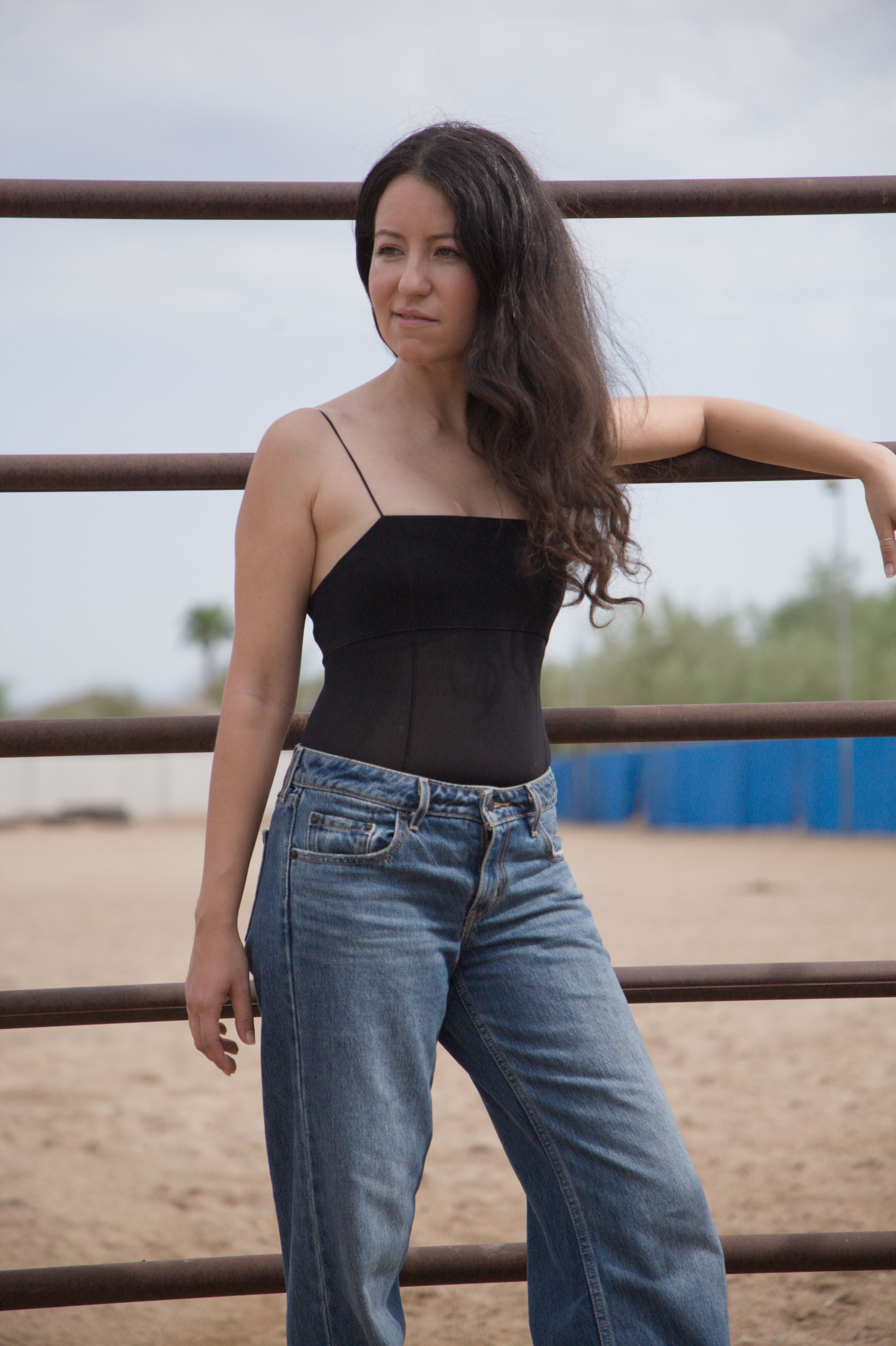 Woman posing casually by outdoor fencing in jeans and top.