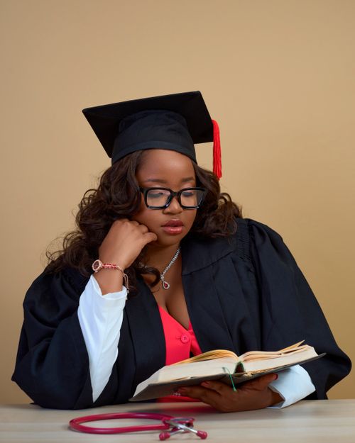 Free Woman in graduation gown reading a book with a stethoscope on the table, symbolizing education and health. Stock Photo