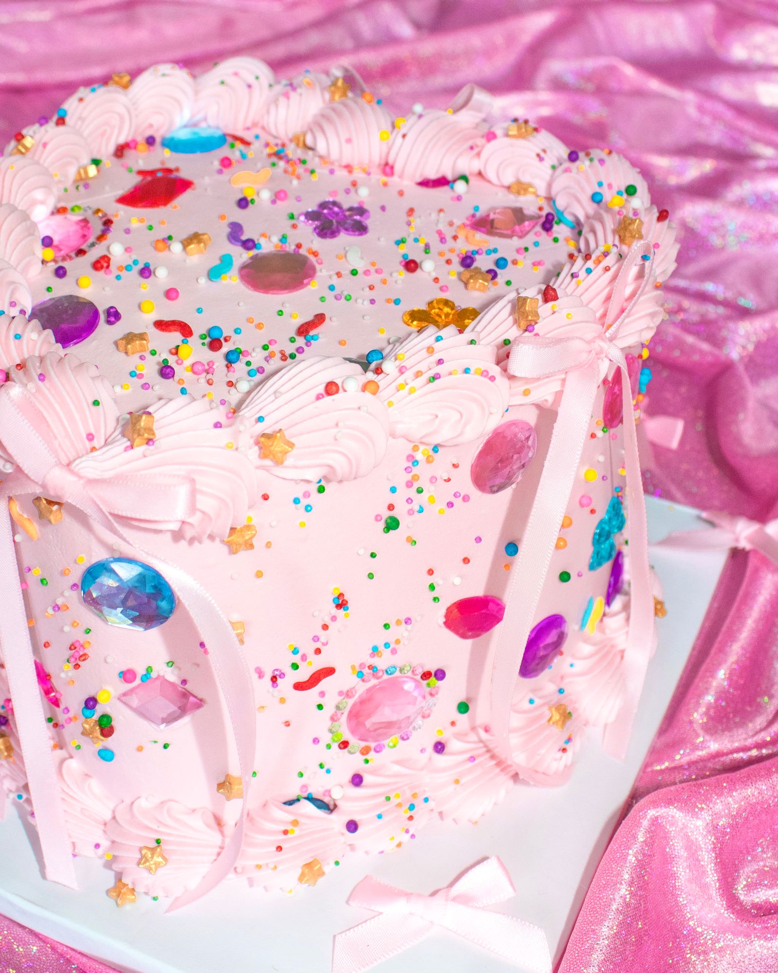 A pink frosted cake covered in oversized colourful gems, sprinkles and ribbon bows on a shiny pink backdrop