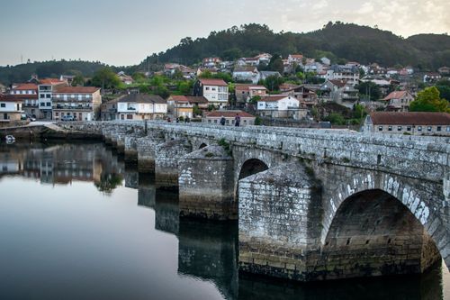 Free A scenic view of a historic stone bridge over a calm river with a picturesque village backdrop. Stock Photo