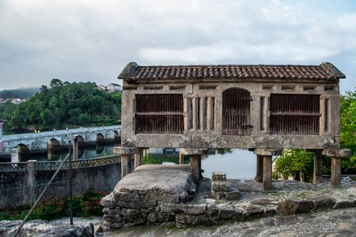 Free Traditional hórreo by a scenic river with lush greenery and a charming stone bridge. Stock Photo