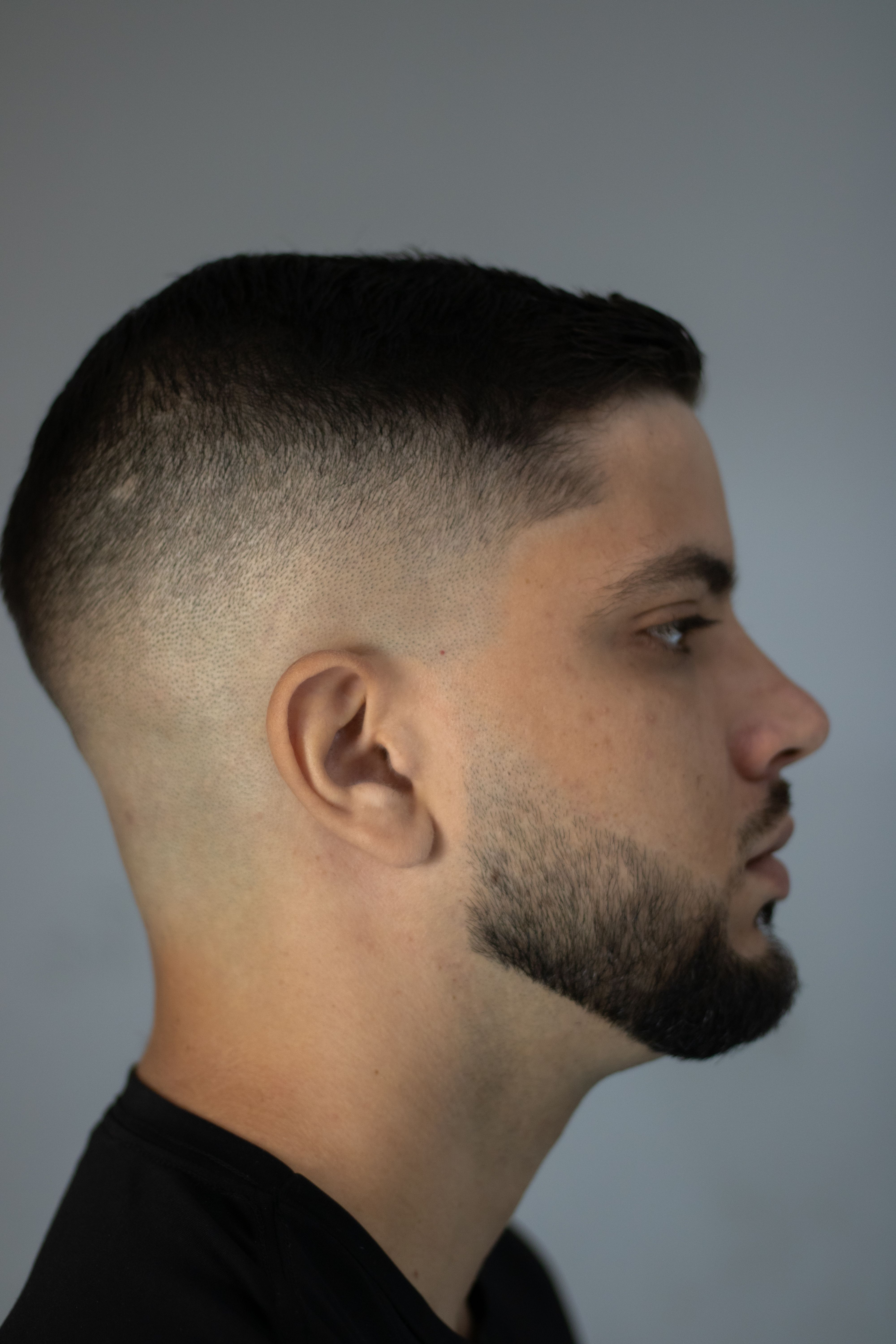 Profile view of young adult male with modern buzz cut and beard on gray background.