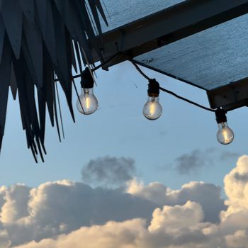 Three hanging bulbs under a roof with a serene cloudy sky in the background at sunset.（イメージ）