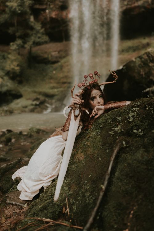 Free A woman dressed in fantasy clothing poses with a sword near a waterfall, set in a mystical forest backdrop. Stock Photo