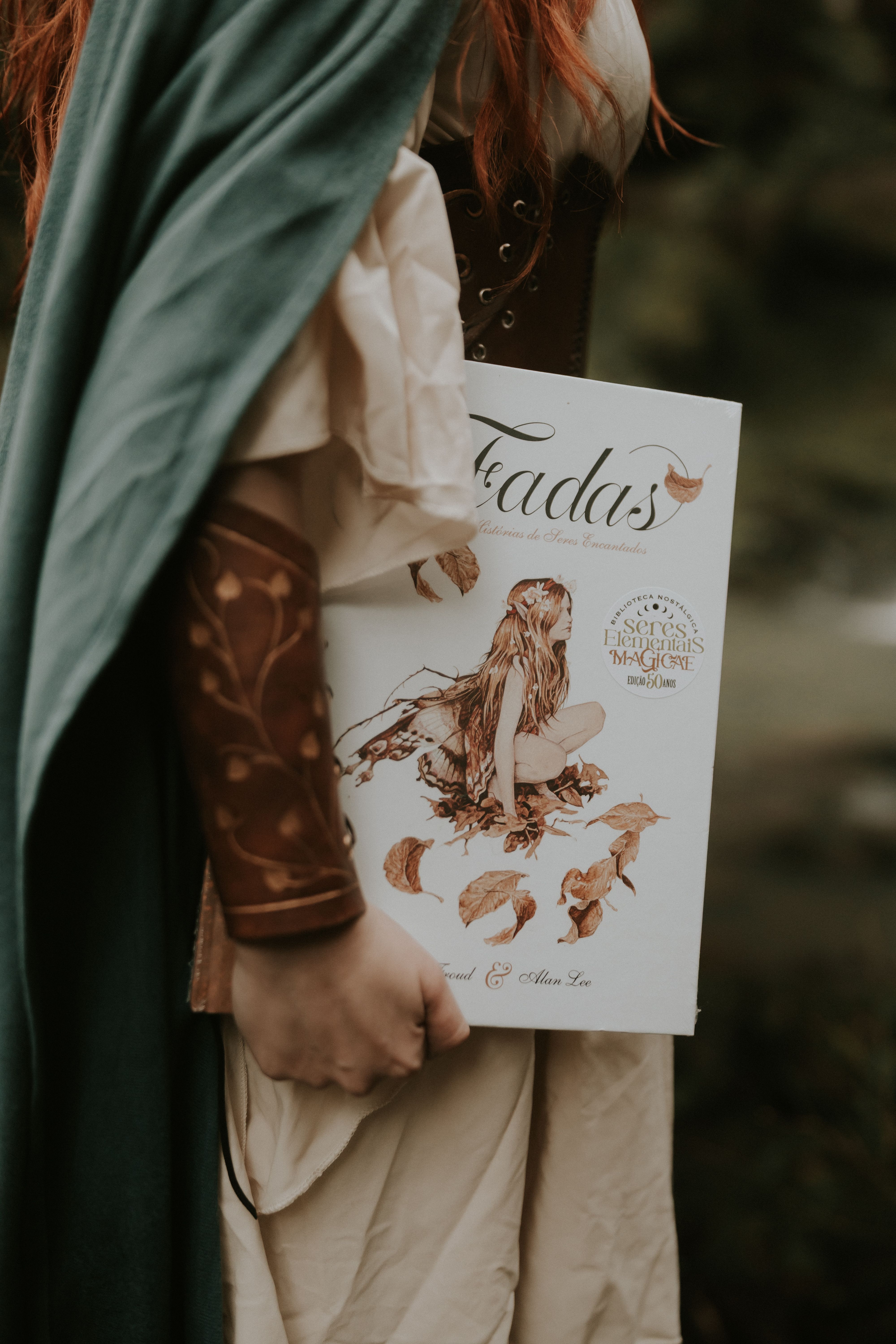 A person dressed in medieval fantasy attire holding a book titled "Fadas" outdoors.