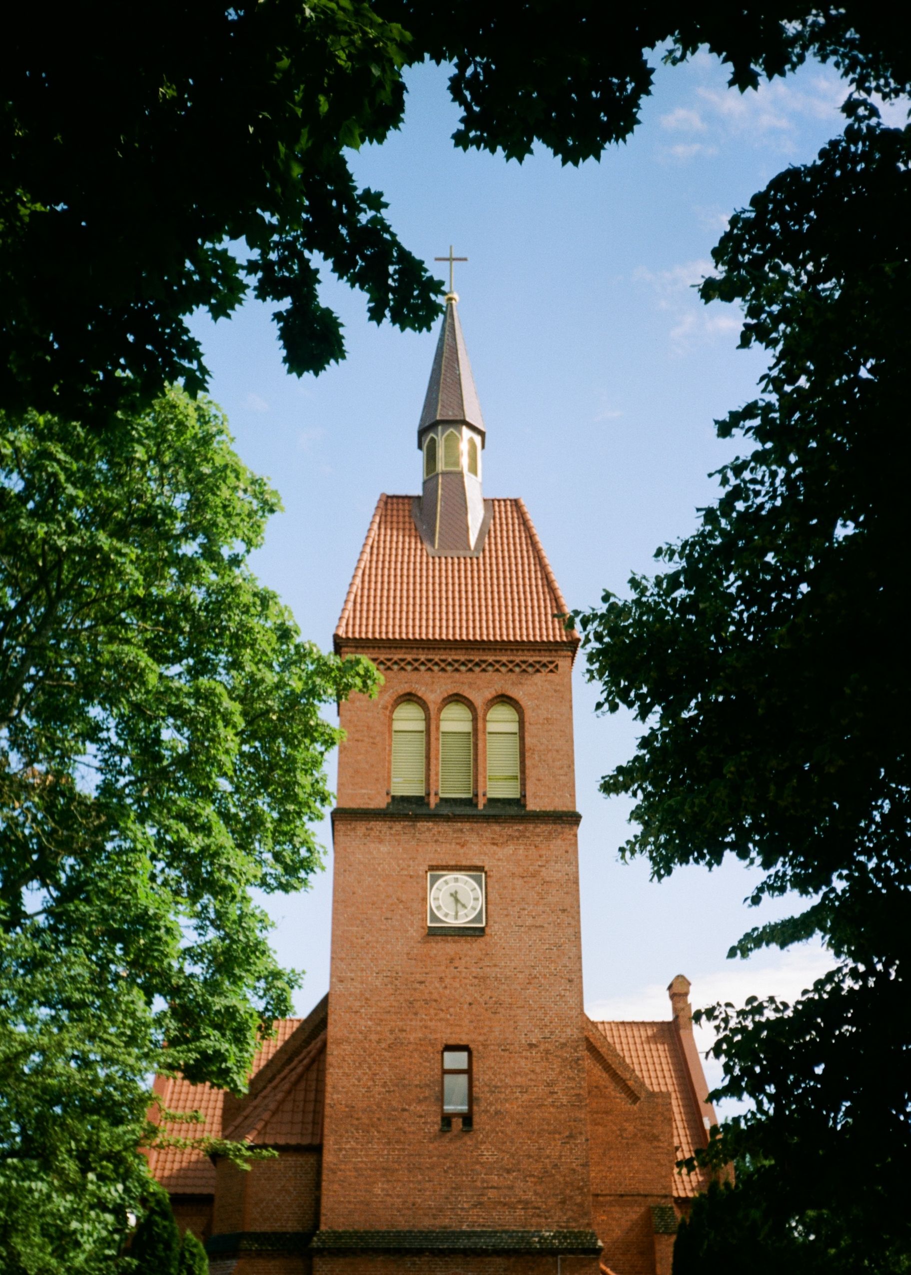 A gothic-style church tower with a cross in a lush, green setting under a clear blue sky.