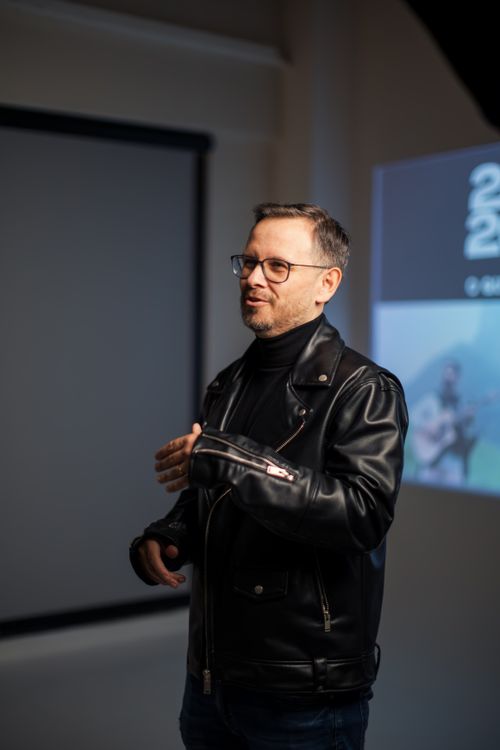 Free Man in a leather jacket and glasses speaking during an indoor presentation. Stock Photo