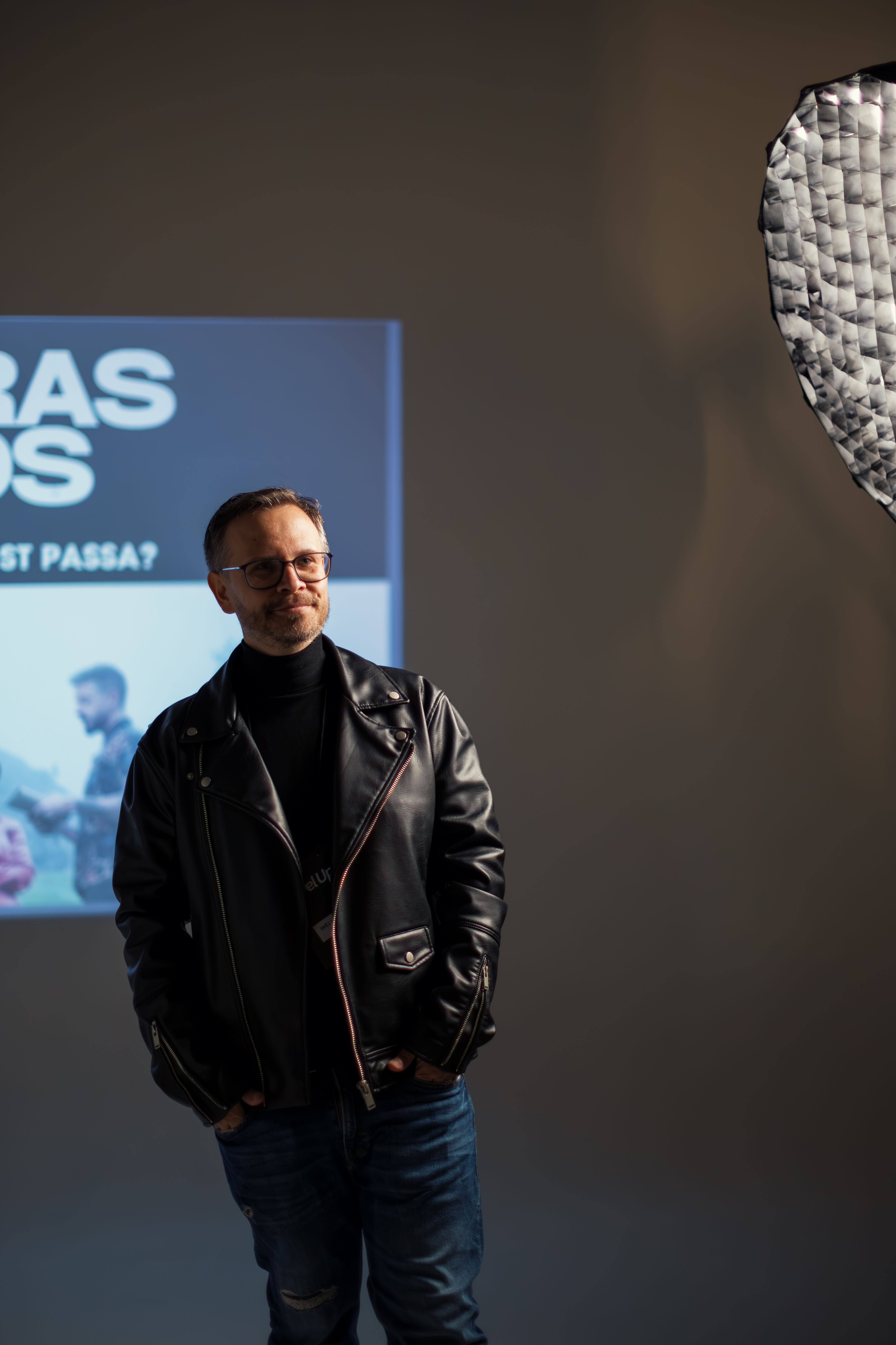 Stylish man wearing a leather jacket and glasses poses in a studio with soft lighting.