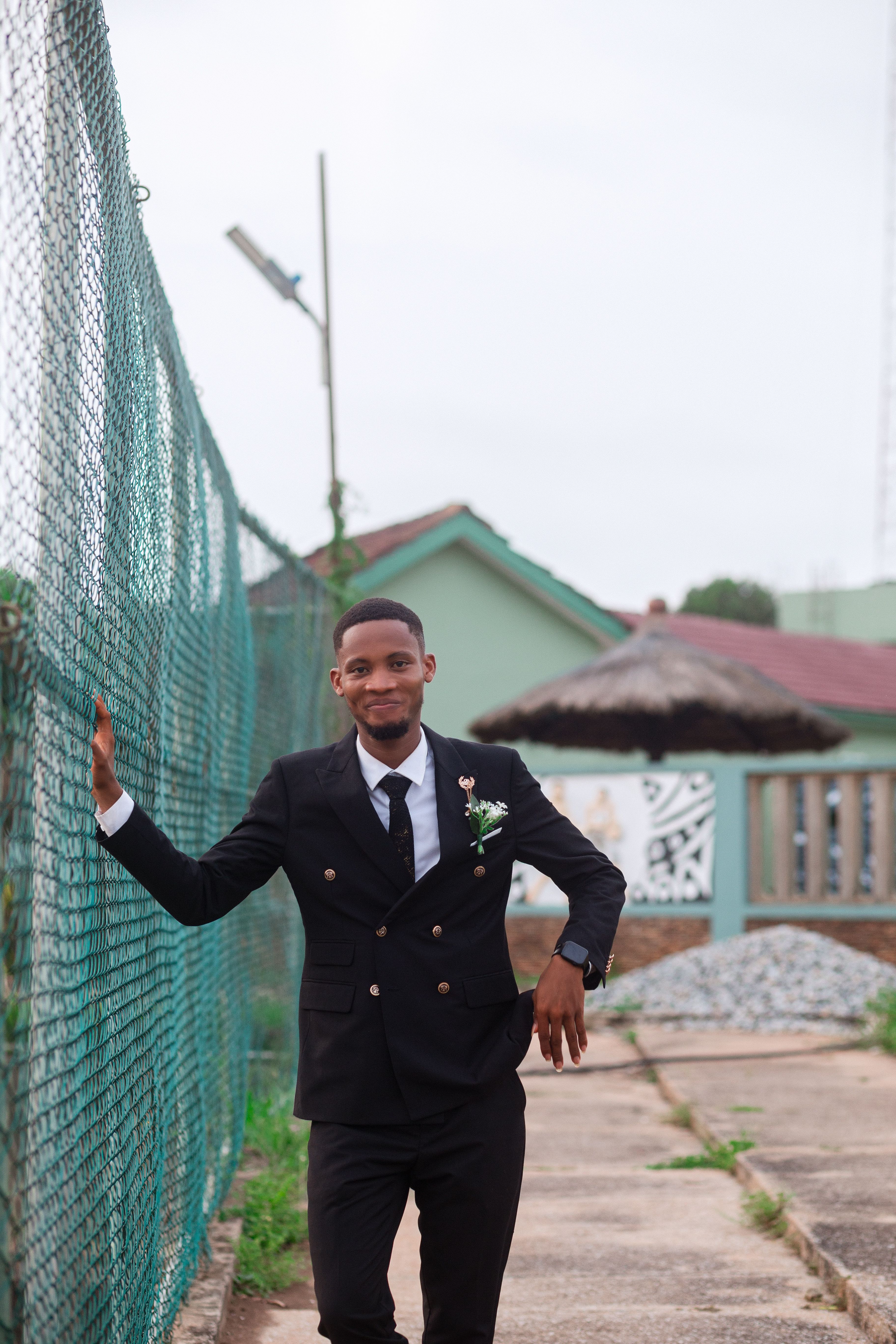 A stylish young man poses confidently in a black suit against a sports field fence.