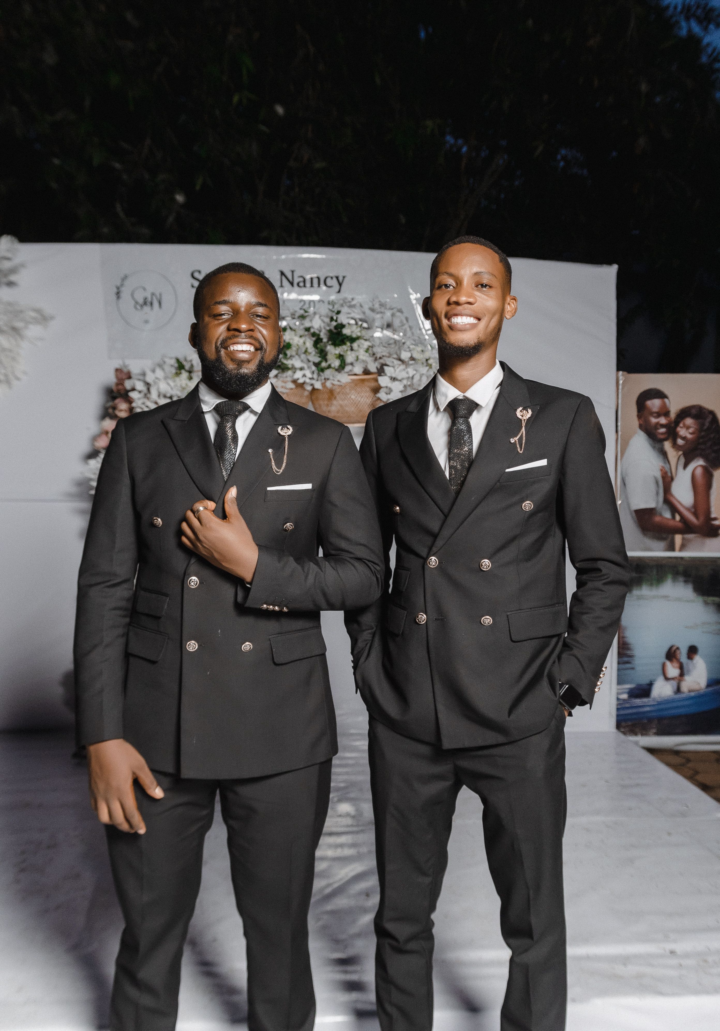 Two men in elegant black suits smiling at an outdoor formal event.