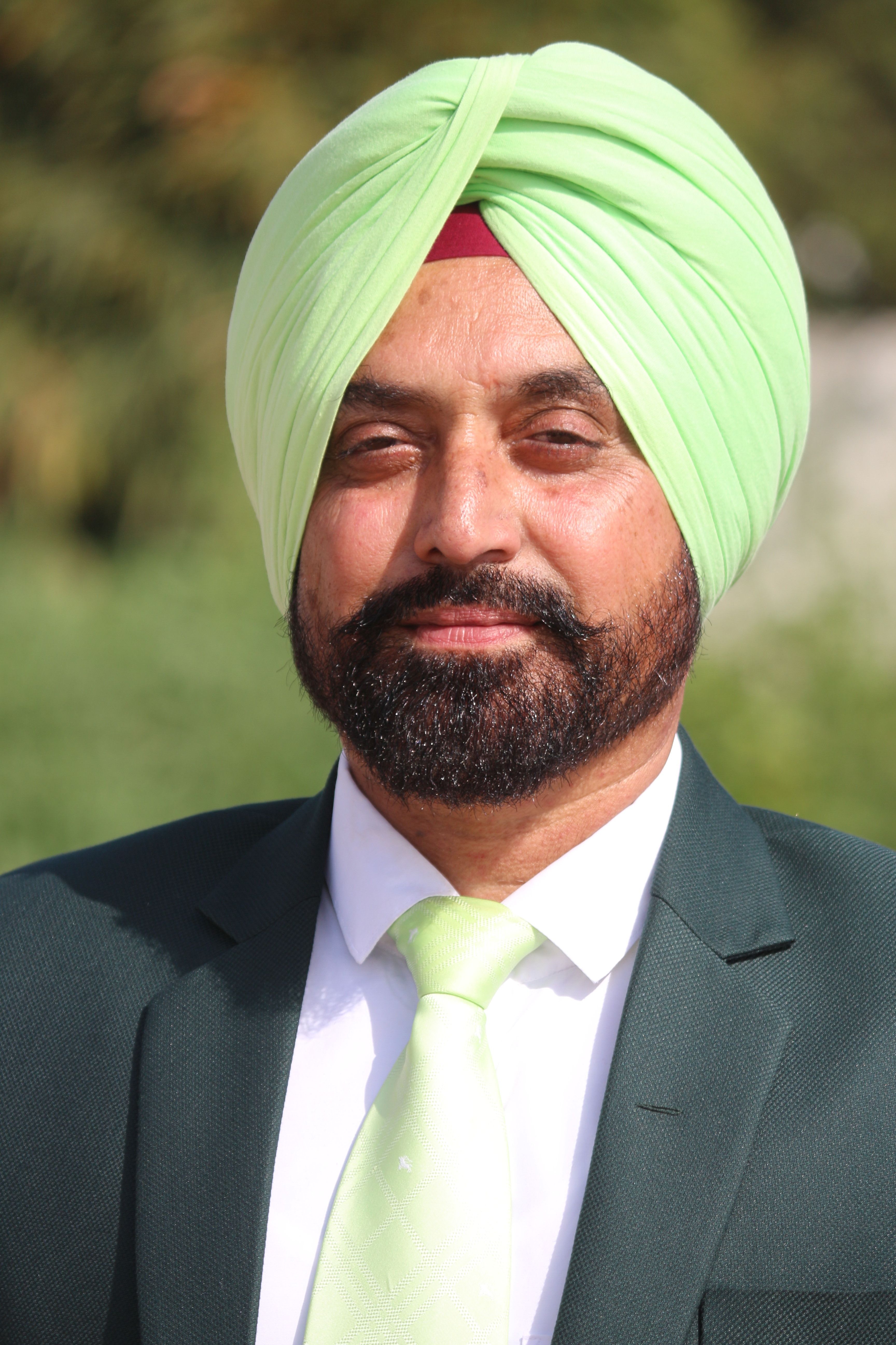 A man wearing a green turban and suit smiling confidently outdoors under bright sunlight.