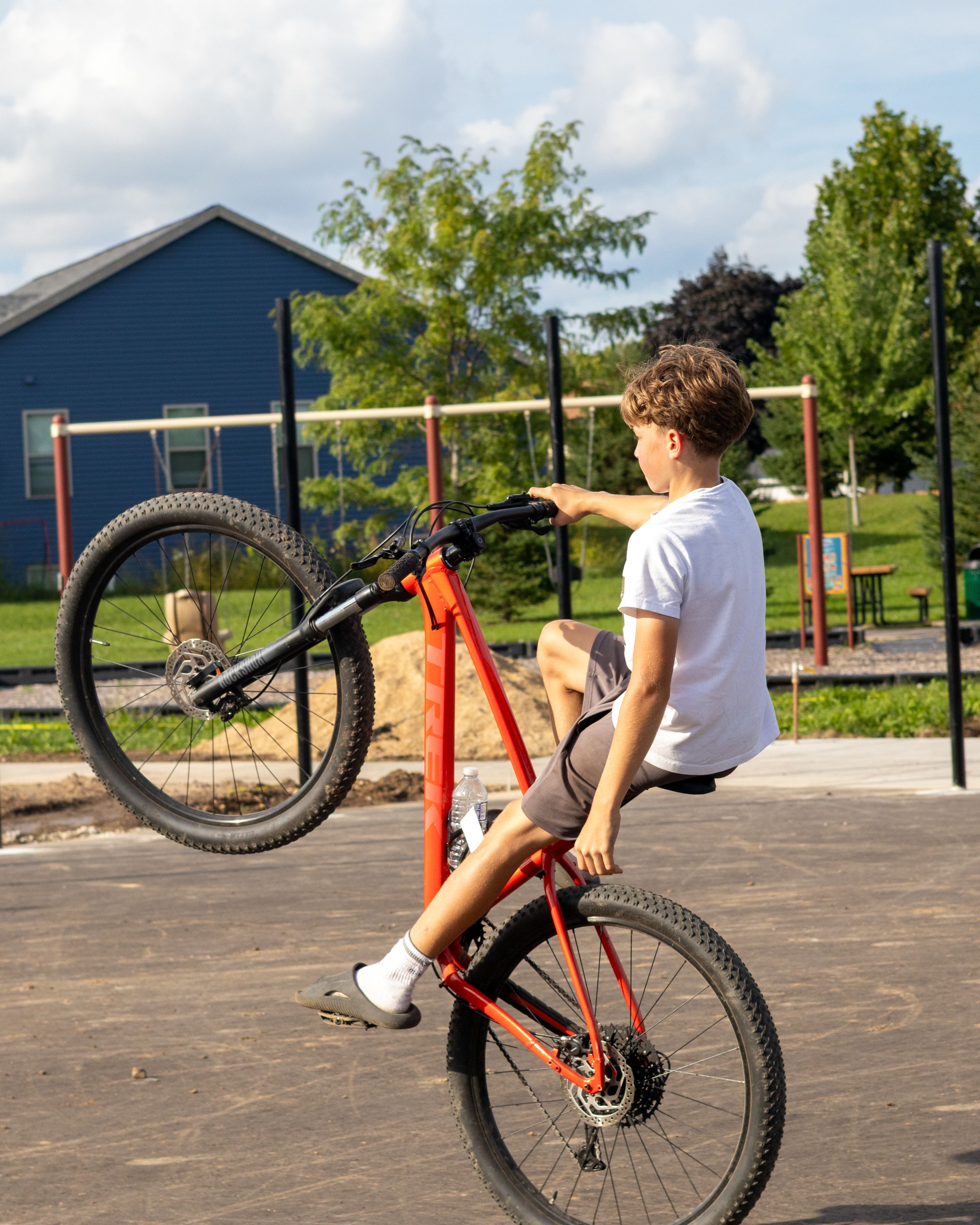 Young boy performing a wheelie on a mountain bike in a sunny park setting, showcasing skill and balance.