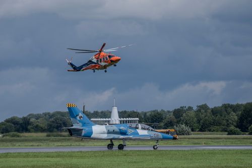 Free A striking scene of a helicopter flying above a stationary fighter jet on a runway with a cloudy sky backdrop. Stock Photo