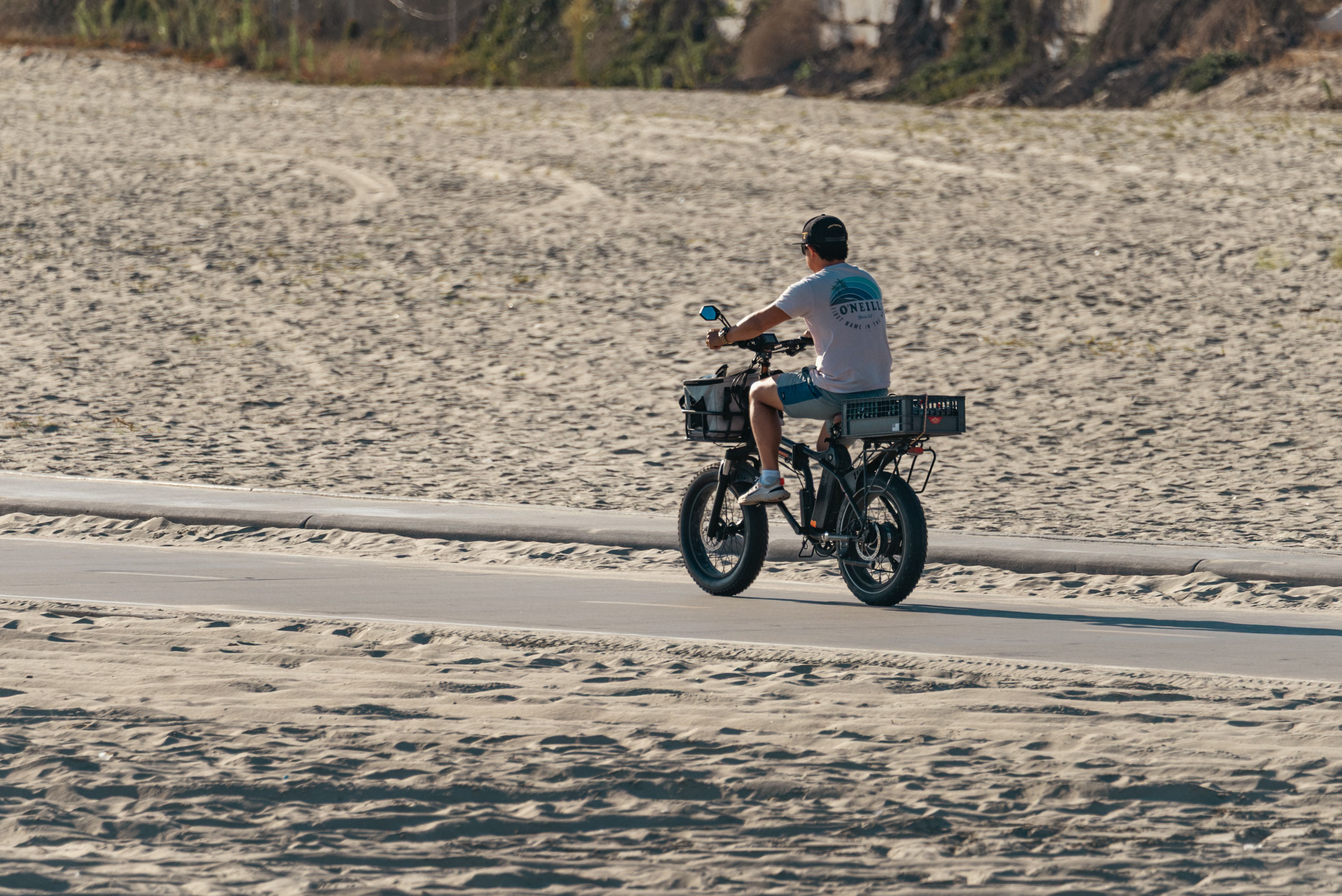 Man riding an electric bike along a sandy beach path under a clear sky.