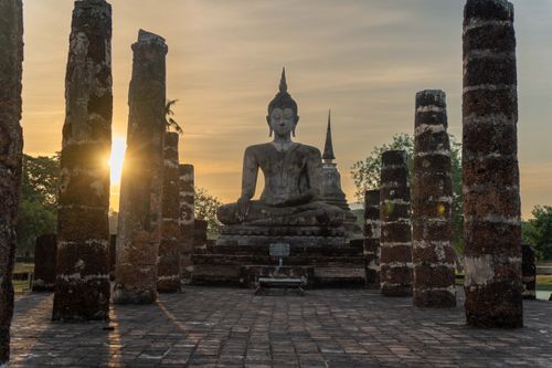Free Serene Buddha statue in Sukhothai Historical Park with sunrise backdrop and ancient pillars. Stock Photo