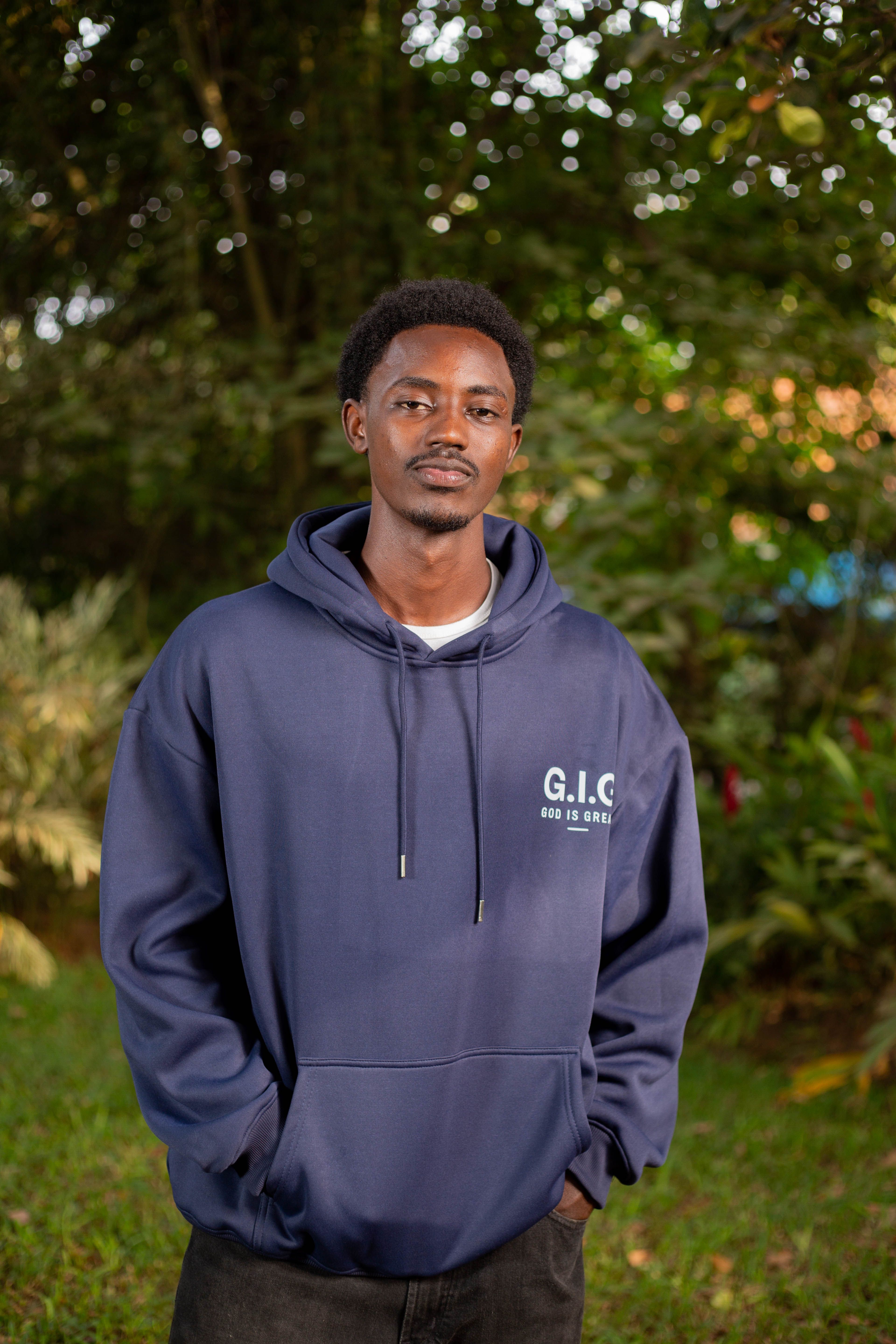 Portrait of a young adult in a blue hoodie standing outdoors with greenery in the background.