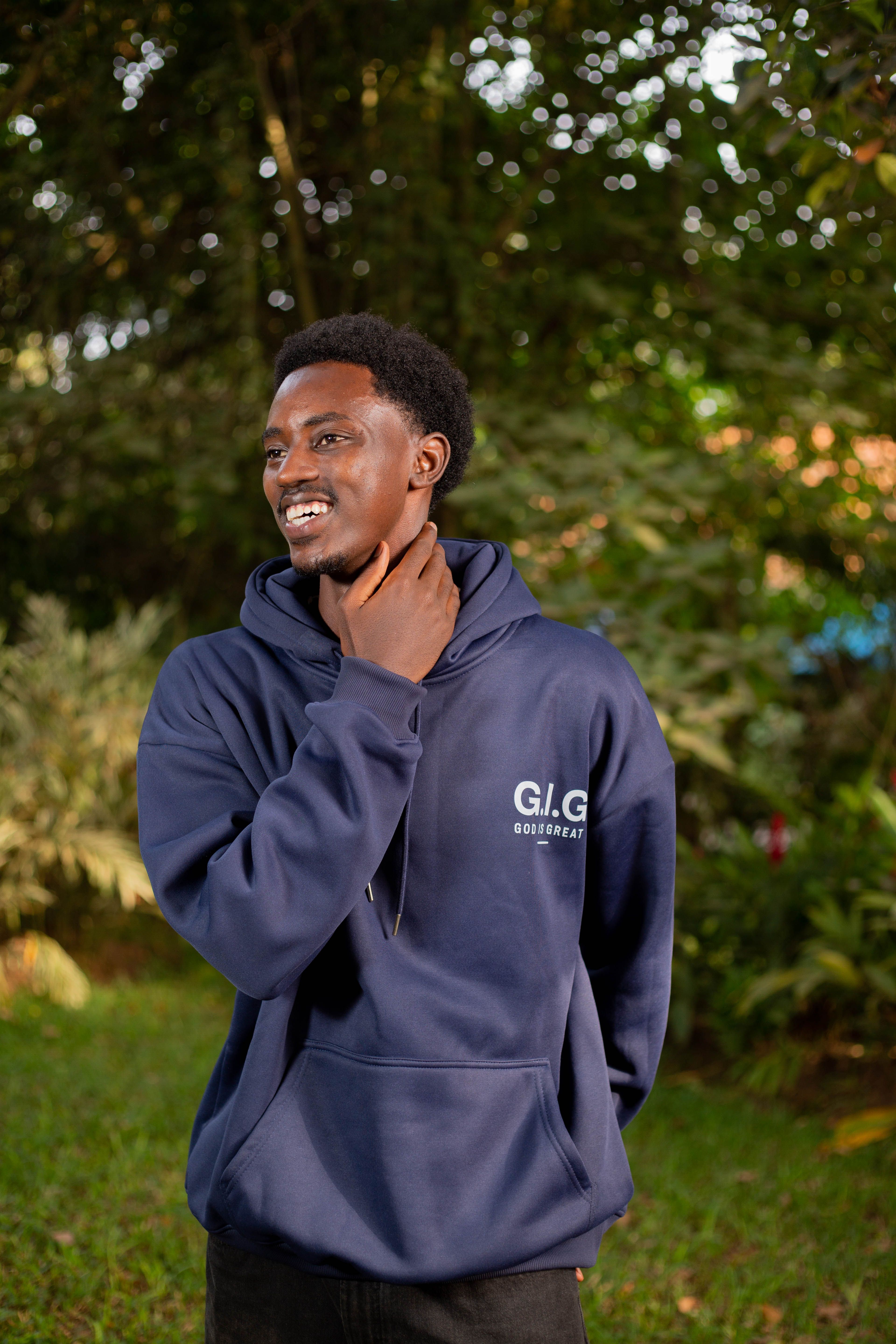 A young man with a blue hoodie smiling in a green outdoor setting.