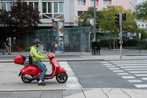Free A person on a red scooter waits at a city crosswalk amid pedestrians and urban buildings. Stock Photo