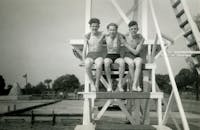 一张老式黑白照片里，三个男孩坐在游泳池边微笑