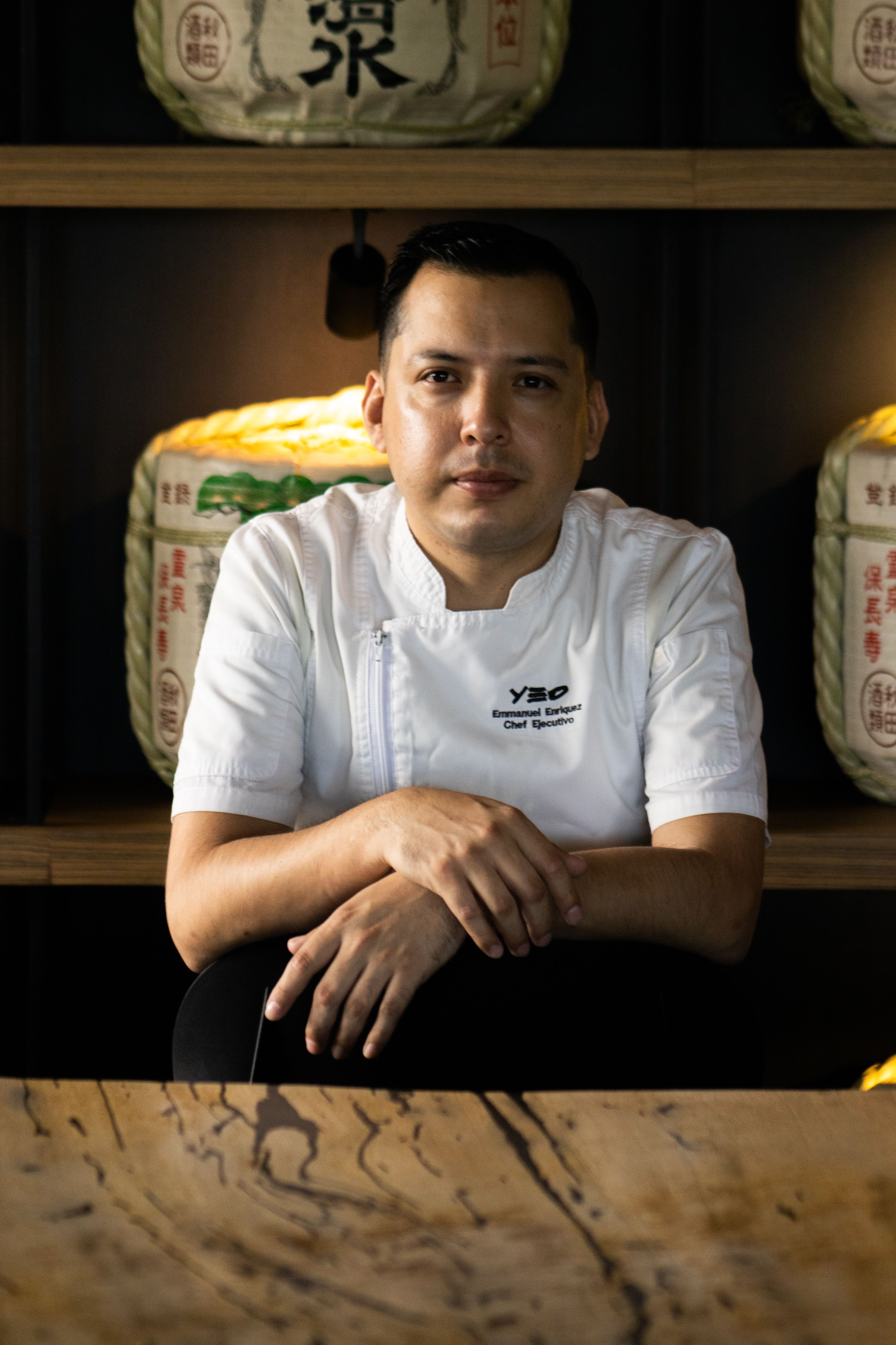 Chef in white uniform seated indoors with Japanese decor.