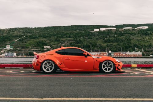 Free Side view of an orange sports car parked by water, green hills in background. Stock Photo