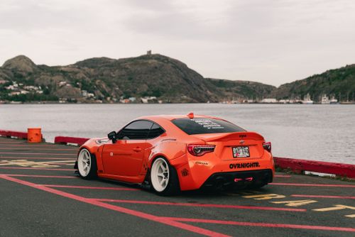 Free Vibrant orange sports car parked by a scenic river with hills in the background, showcasing automotive design. Stock Photo