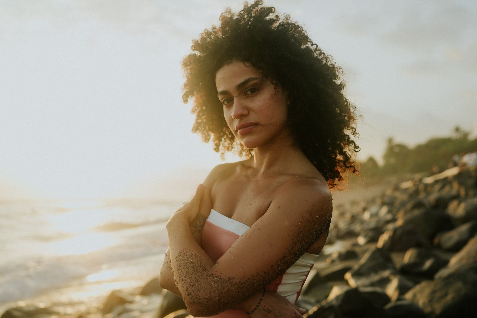 Woman with curly hair and crossed arms stands on a rocky beach at sunset, looking at the camera