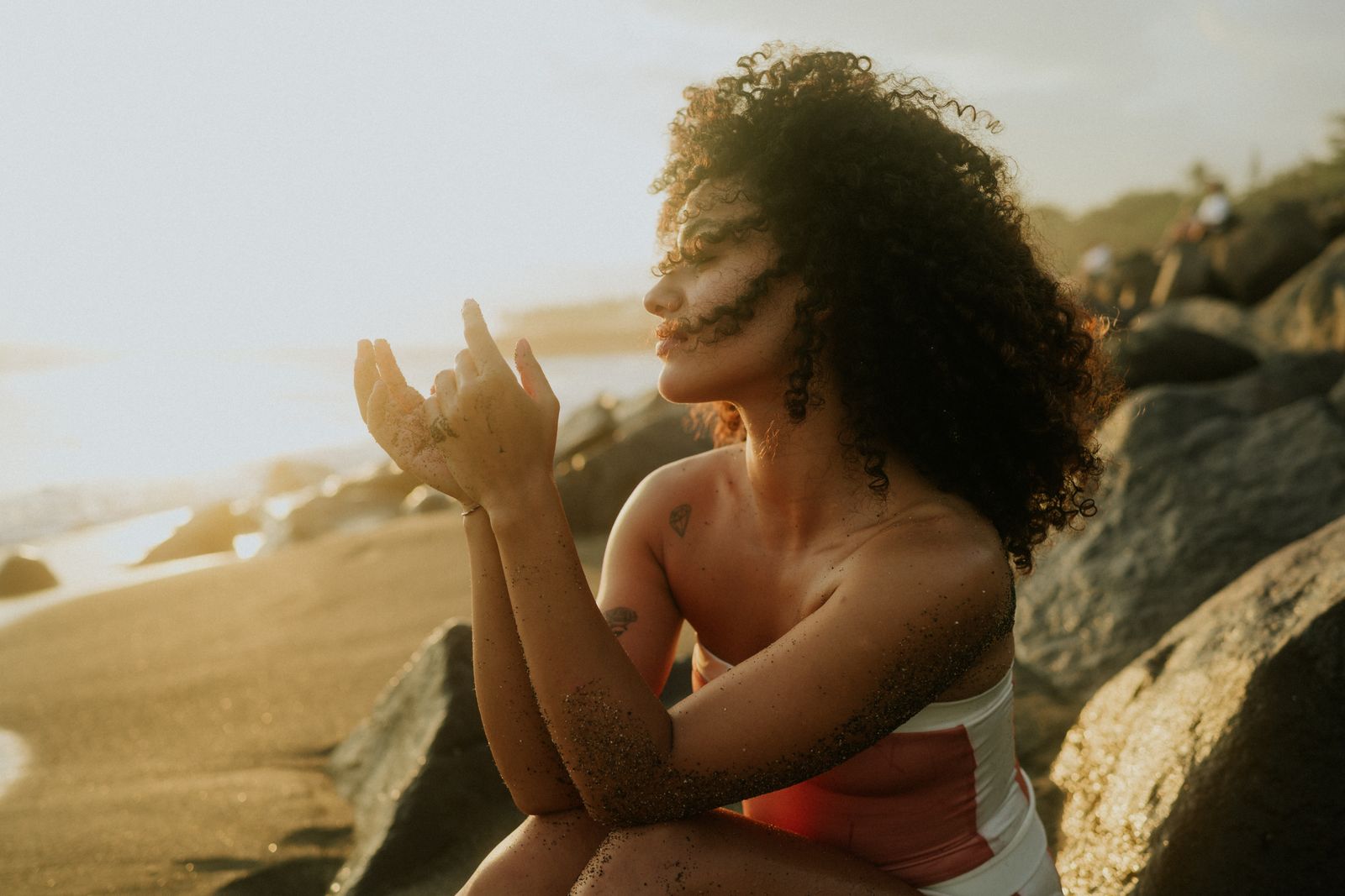 Woman with curly hair sitting on a sunlit beach, hands raised, sand on her arms at golden hour
