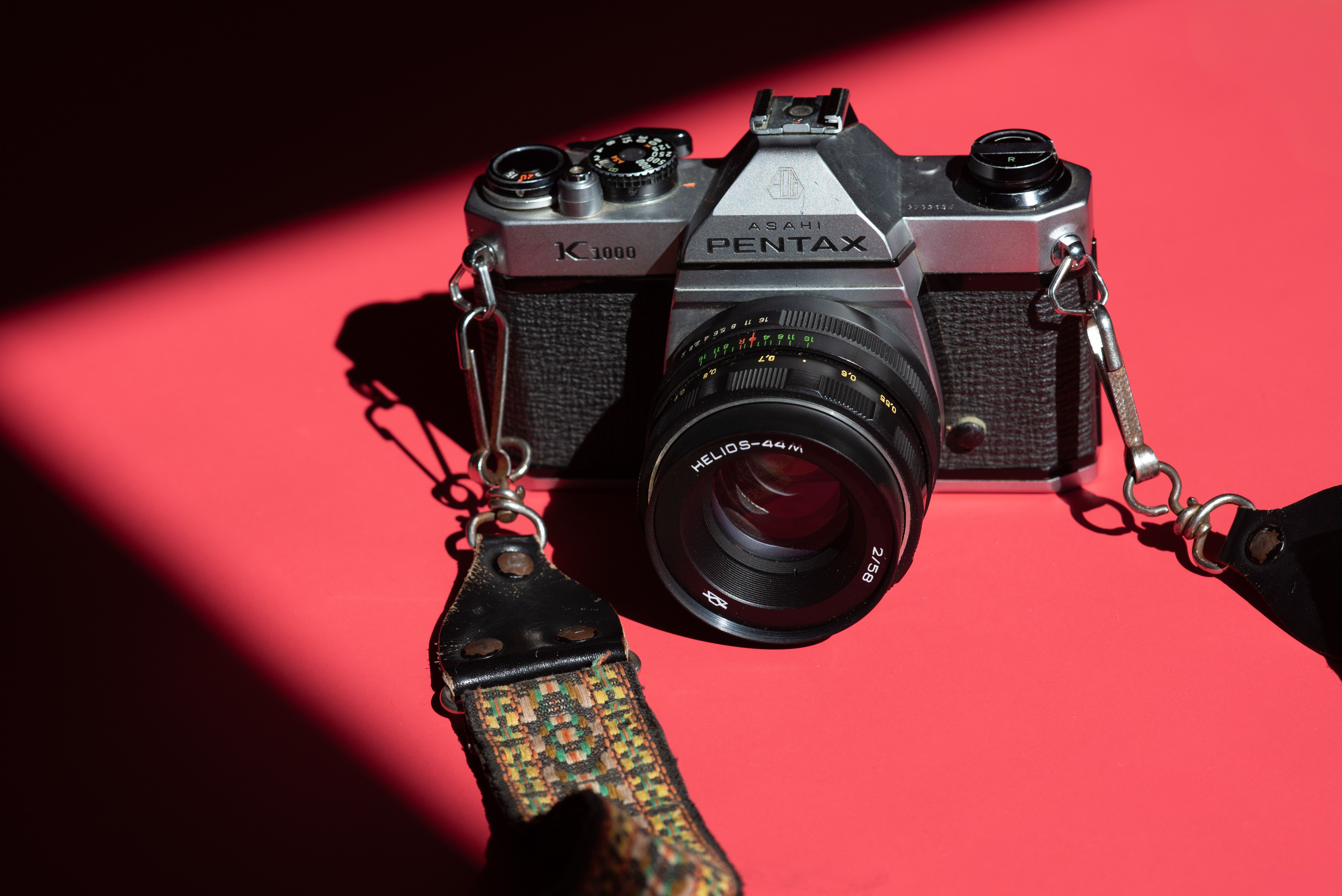A classic Pentax K1000 camera with a Helios-44 lens on a striking red background, casting shadows.