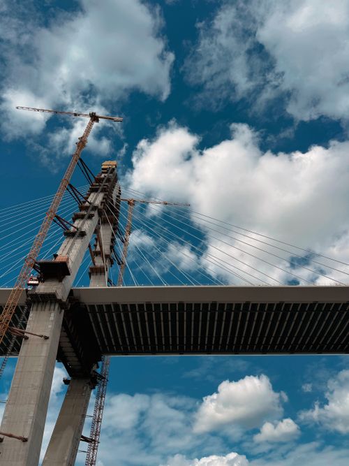 Free Dramatic view of a cable-stayed bridge construction against a cloudy blue sky with cranes. Stock Photo