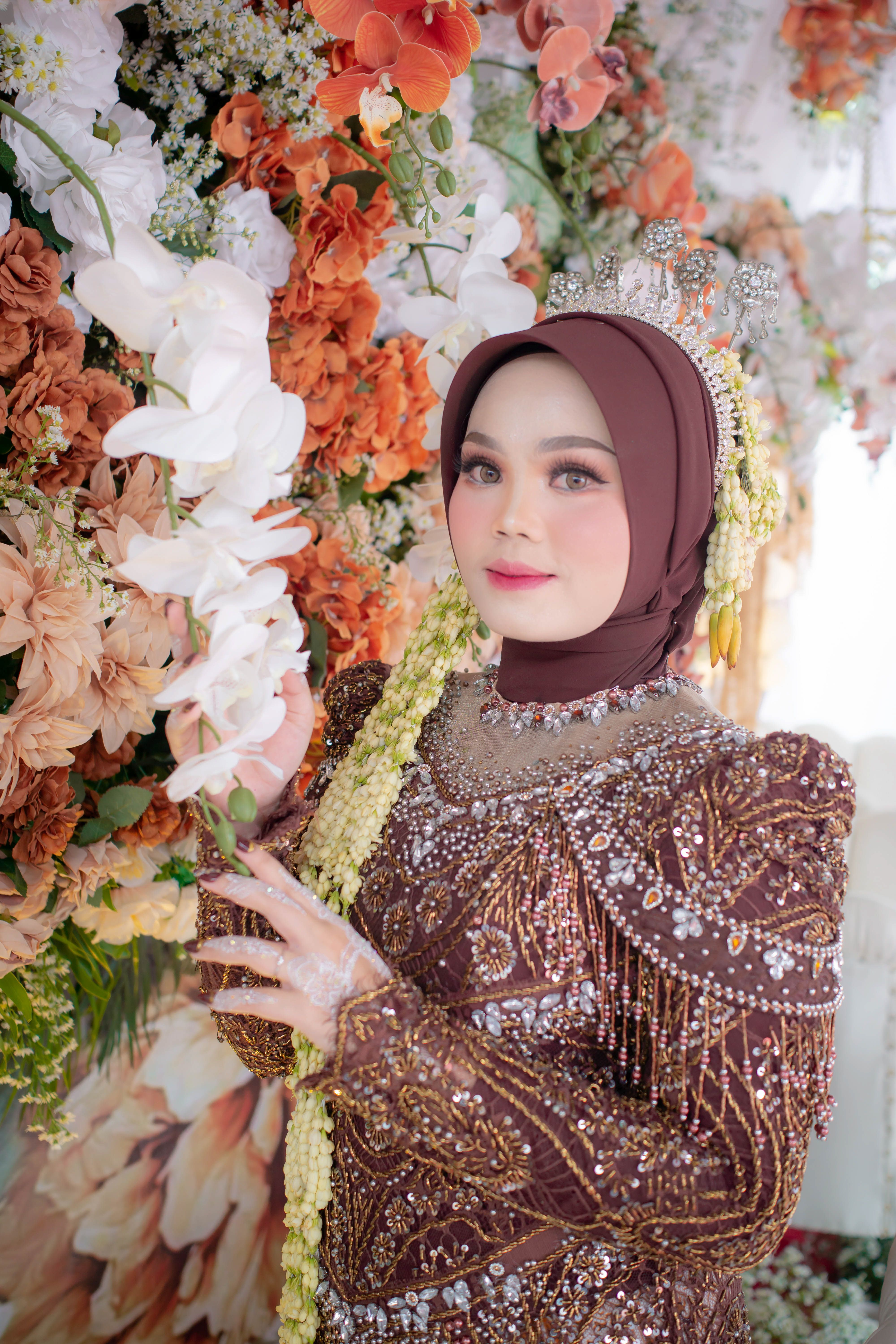 Bride in ornate dress and hijab surrounded by vibrant floral arrangement.