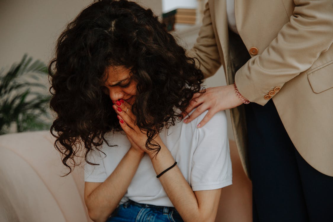 A therapist stands by her upset patient, who is crying with her hands by her face, and comforts her.