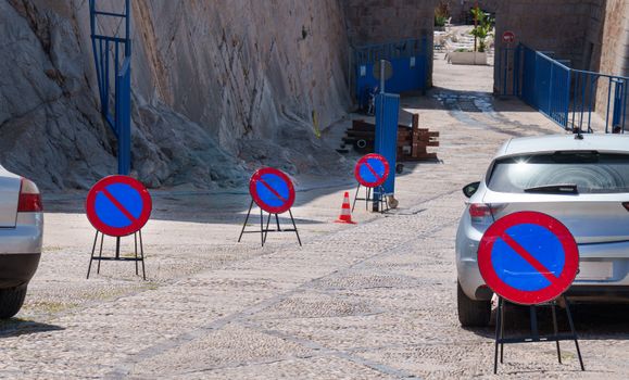 A row of no parking signs on a cobblestone street with parked cars, emphasizing road restrictions.