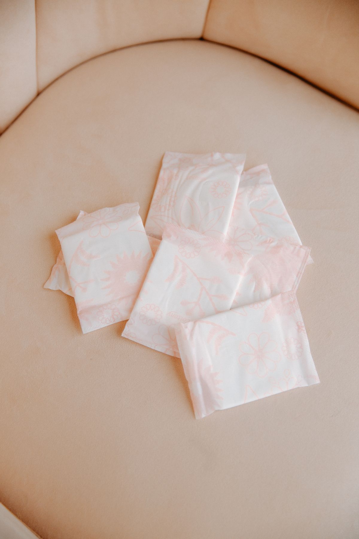 Several individually wrapped sanitary pads with a pale floral pattern resting on a cream velvet chair