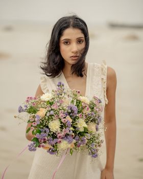 A young woman holding a colorful bouquet on a sandy beach day.