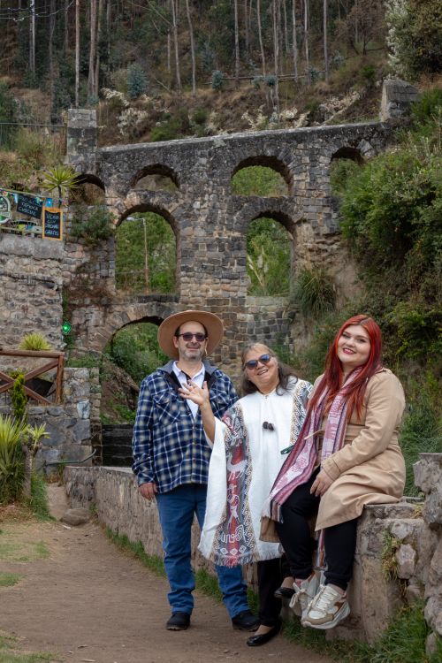 Free Three adults pose in front of a historic stone bridge in a lush outdoor setting. Stock Photo