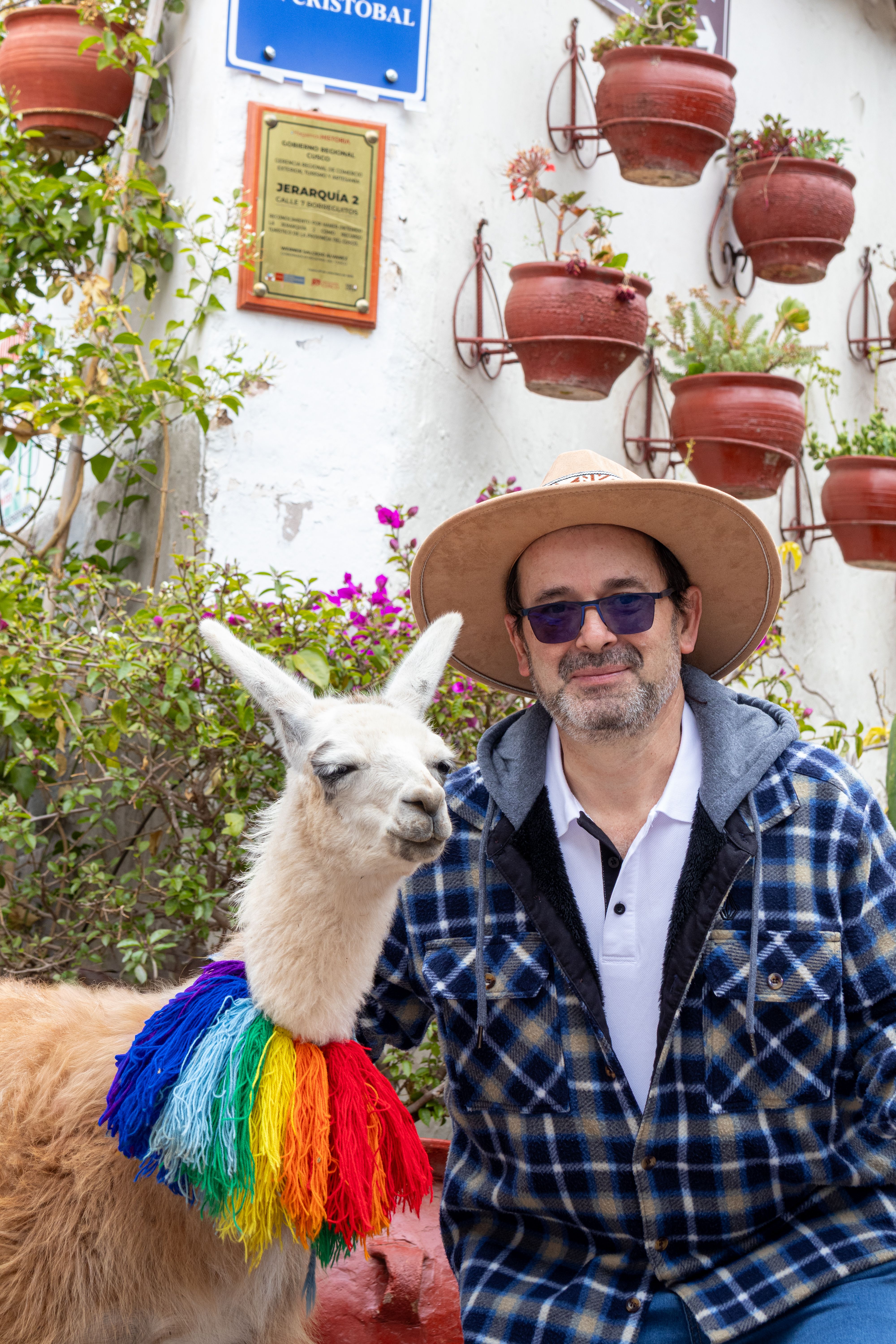 A man wearing a hat poses with a llama in a colorful poncho against an outdoor wall.