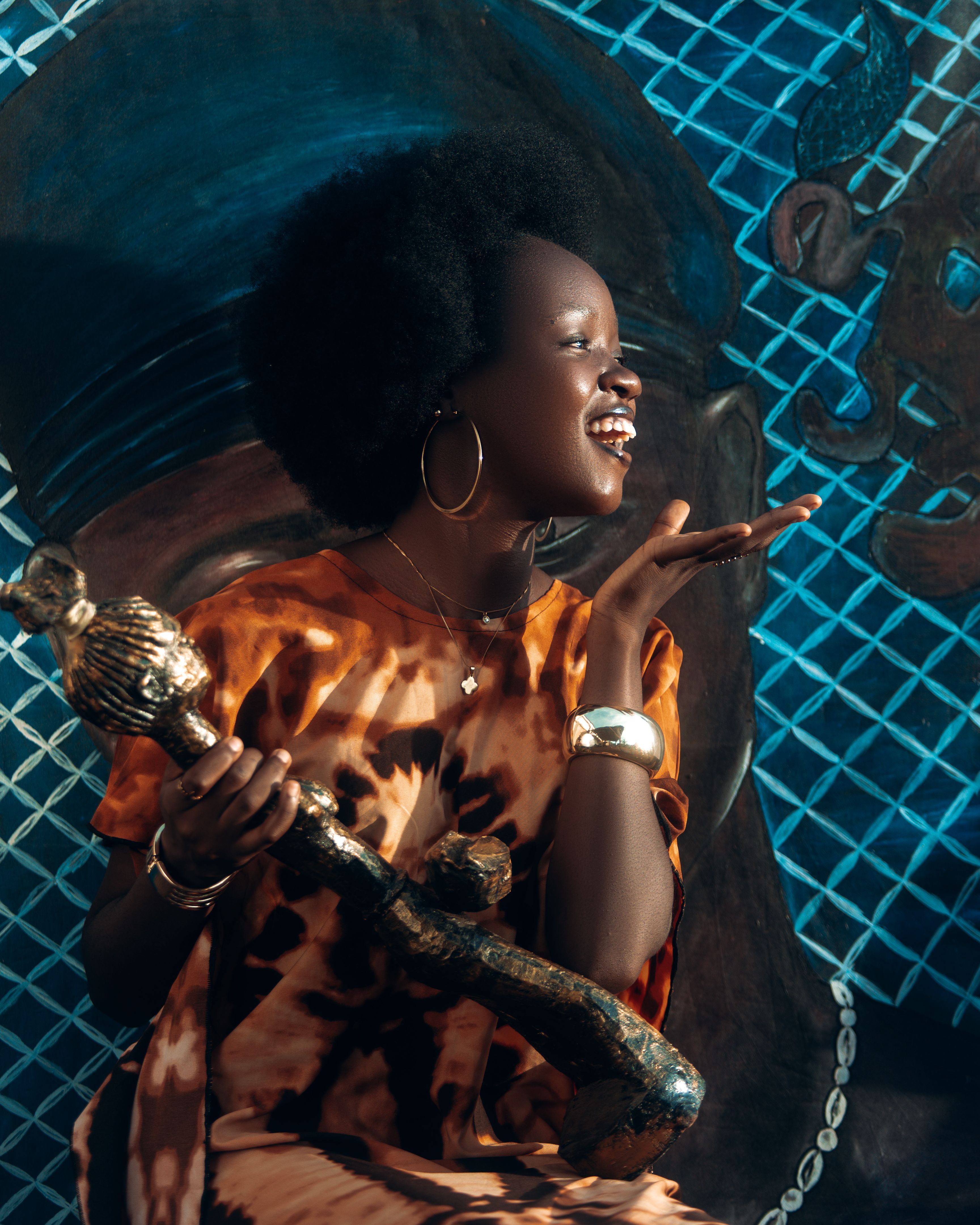 A smiling woman in vibrant clothing holds a unique decorative object against a colorful mural background.
