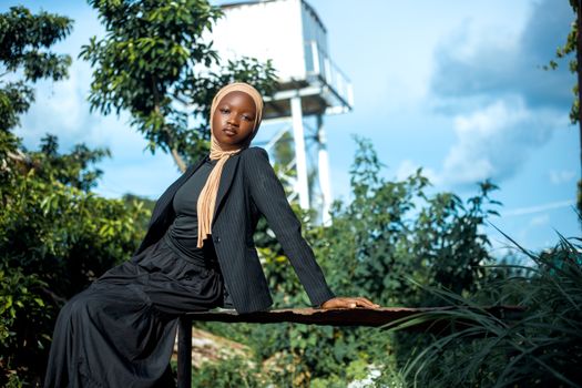 Elegant woman in hijab posing outdoors against lush greenery and blue sky.