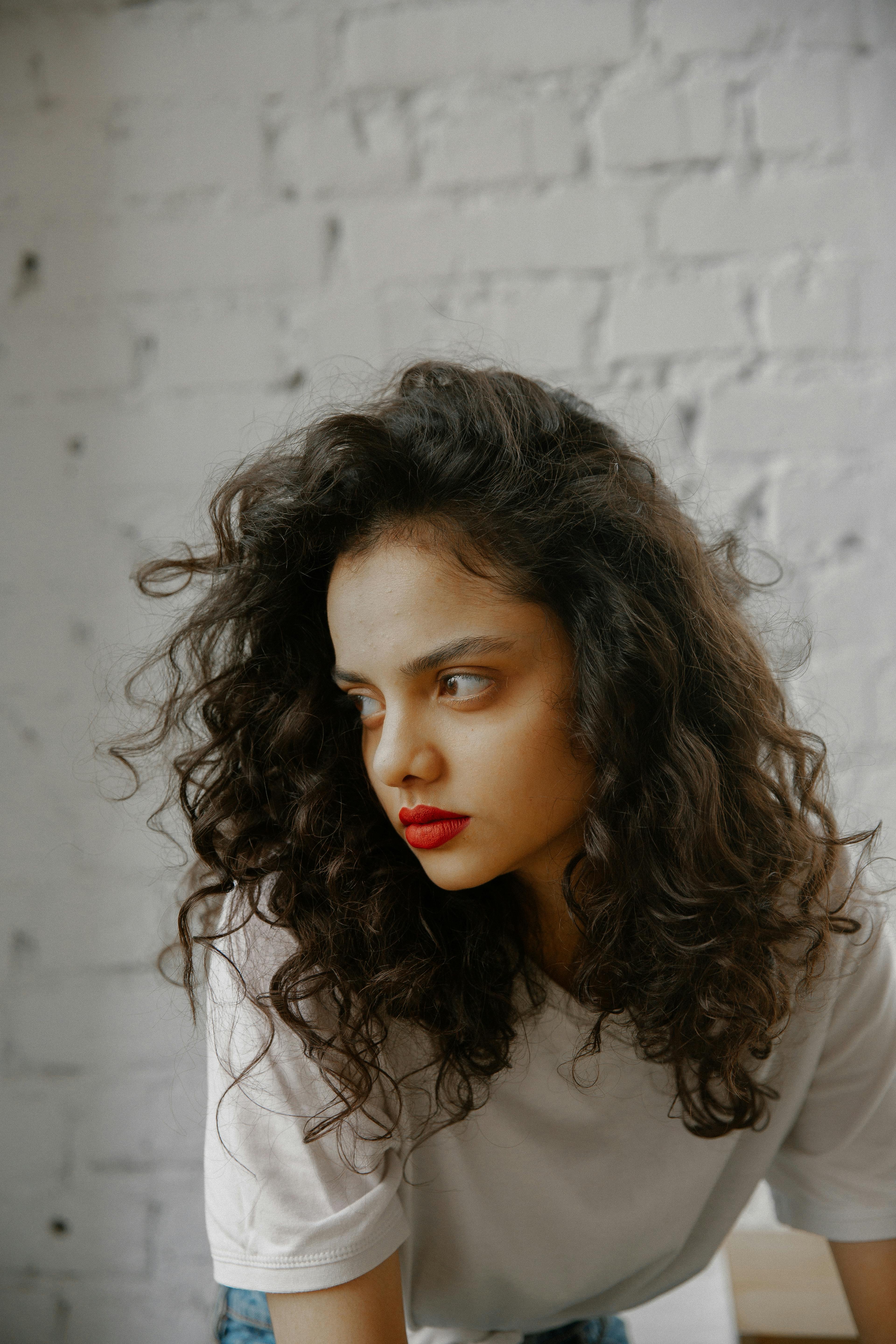 Free A thoughtful brunette woman with curly hair gazes away, set against a textured white brick wall, conveying introspection. Stock Photo