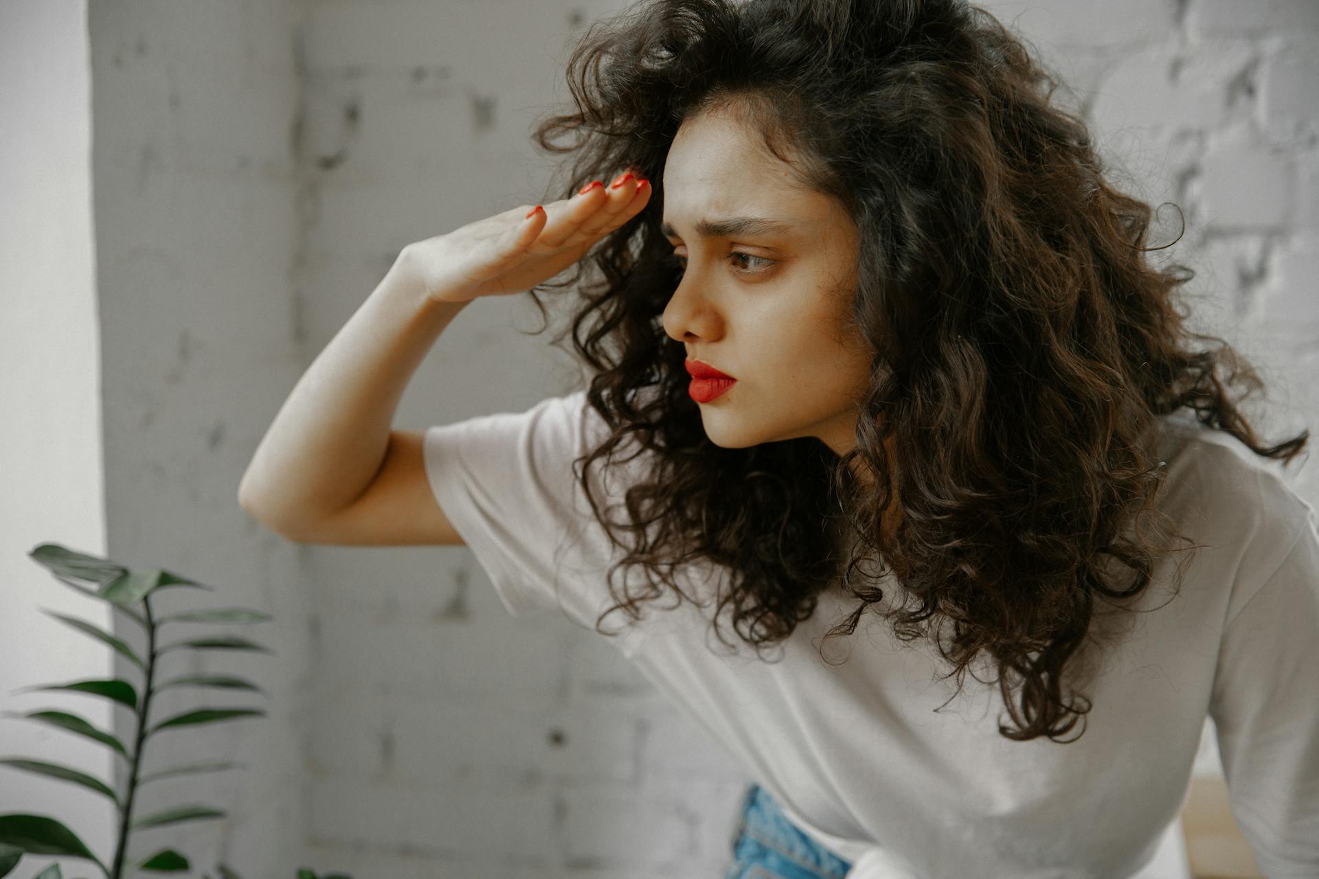 Curly-haired woman in white t-shirt searching indoors with hand above eyes.