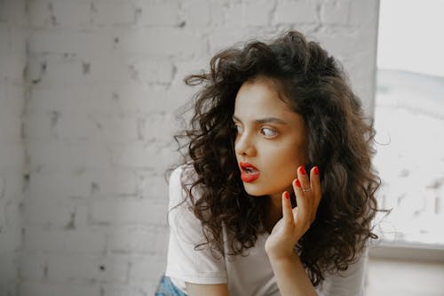 Free Young woman with curly hair and red lipstick looks surprised indoors. Stock Photo