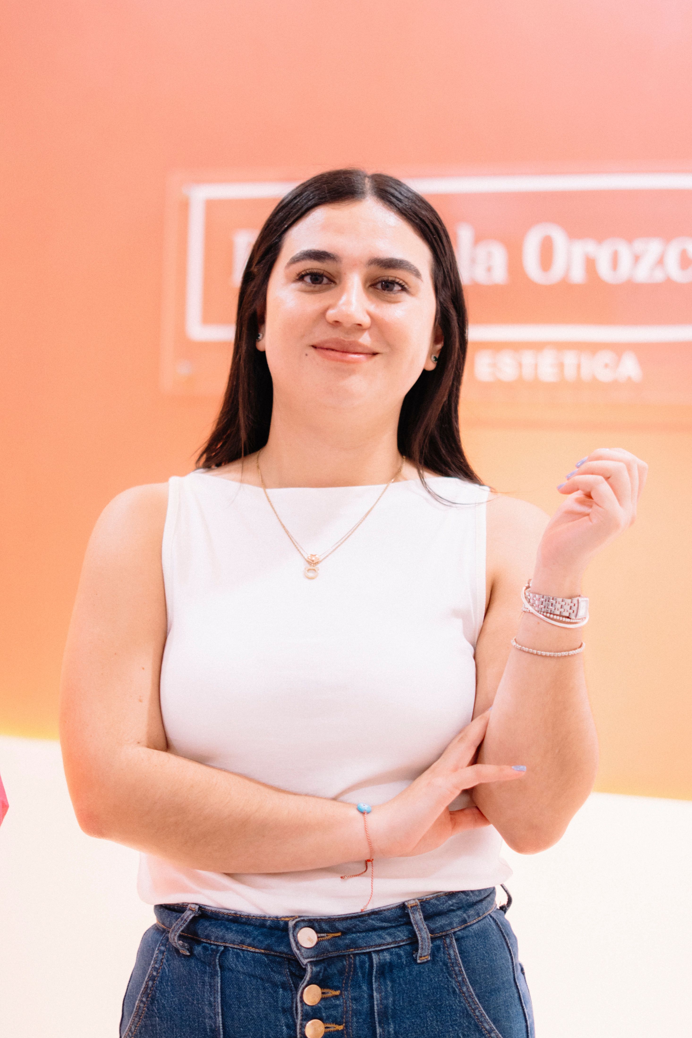 Portrait of a confident young woman in jeans and white top, standing indoors with an orange background.